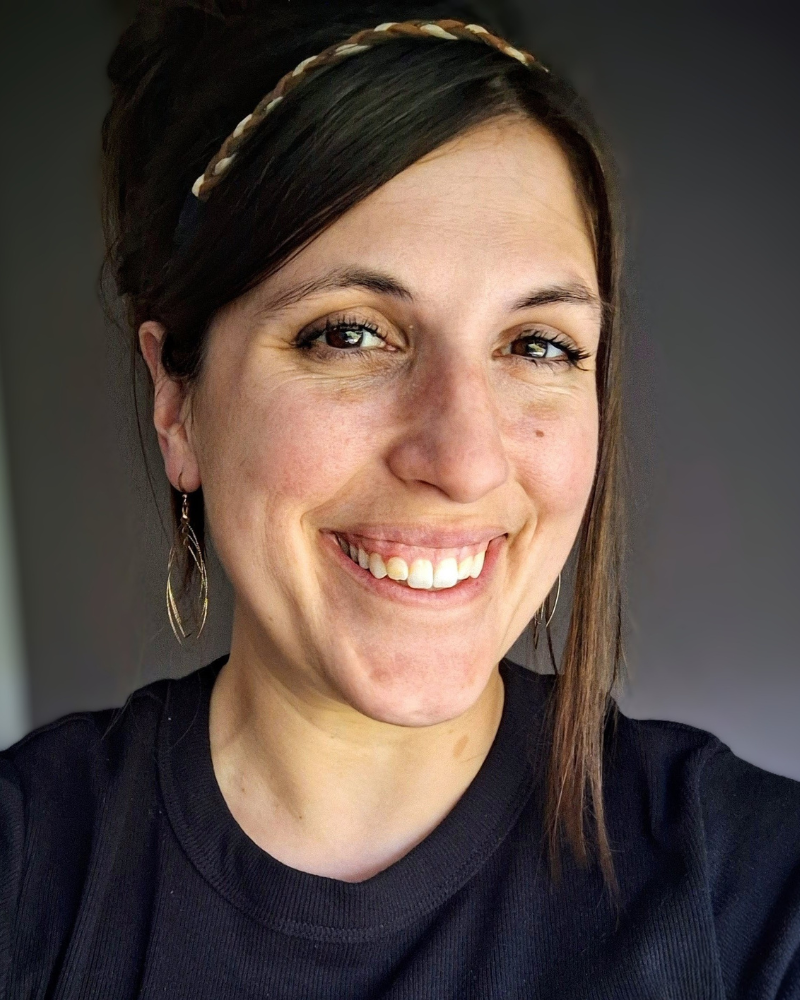 Close-up portrait of a smiling woman with dark hair wearing a braided headband and black top, photographed against a softly lit background.